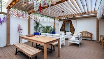 An outdoor patio with a wooden table and benches, surrounded by white cushioned seating and hanging floral decorations.