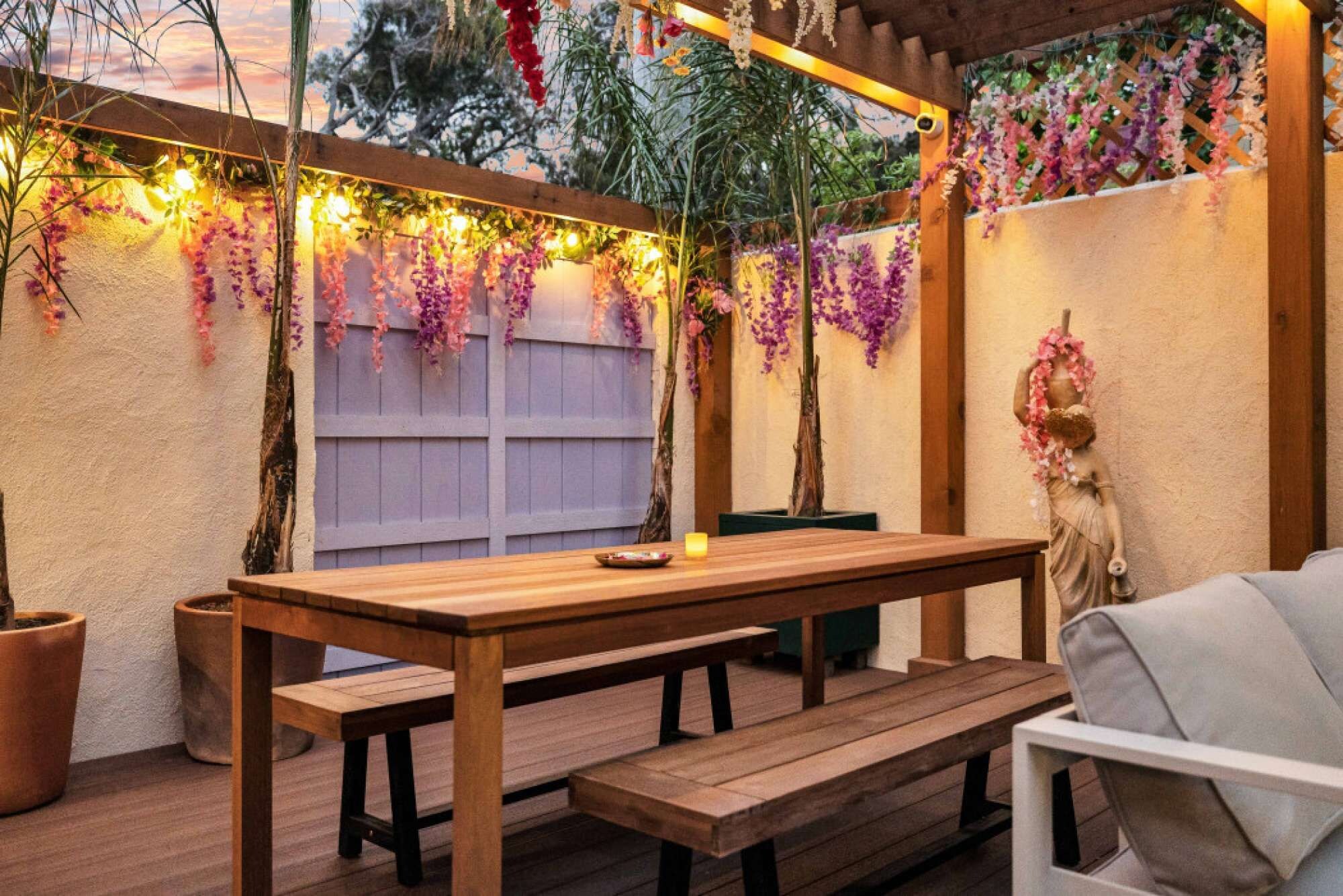 An outdoor patio with a wooden table and benches, surrounded by potted plants and a decorative statue.