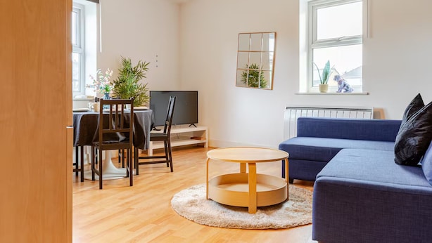 Una sala de estar moderna con un sofá azul, una mesa de centro redonda de madera, un televisor y una zona de comedor con mesa y sillas.