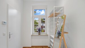 Een kamer met een houten vloer, een raam met uitzicht op gebouwen, een wit plank met spullen, en een witte deur.