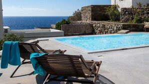 A poolside area with two wooden lounge chairs, a stone wall, and a view of the ocean. - Ormos Seaside Villa G With Private Pool (Tinos)