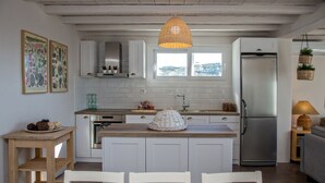 A modern kitchen with white cabinets, a stainless steel refrigerator, a wooden dining table with chairs, and a hanging wicker light fixture. - Ormos Seaside Villa G With Private Pool (Tinos)