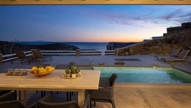 Ein Außenpoolbereich mit Blick auf das Meer bei Sonnenuntergang, umgeben von Liegestühlen und einem Esstisch mit Obstschale.