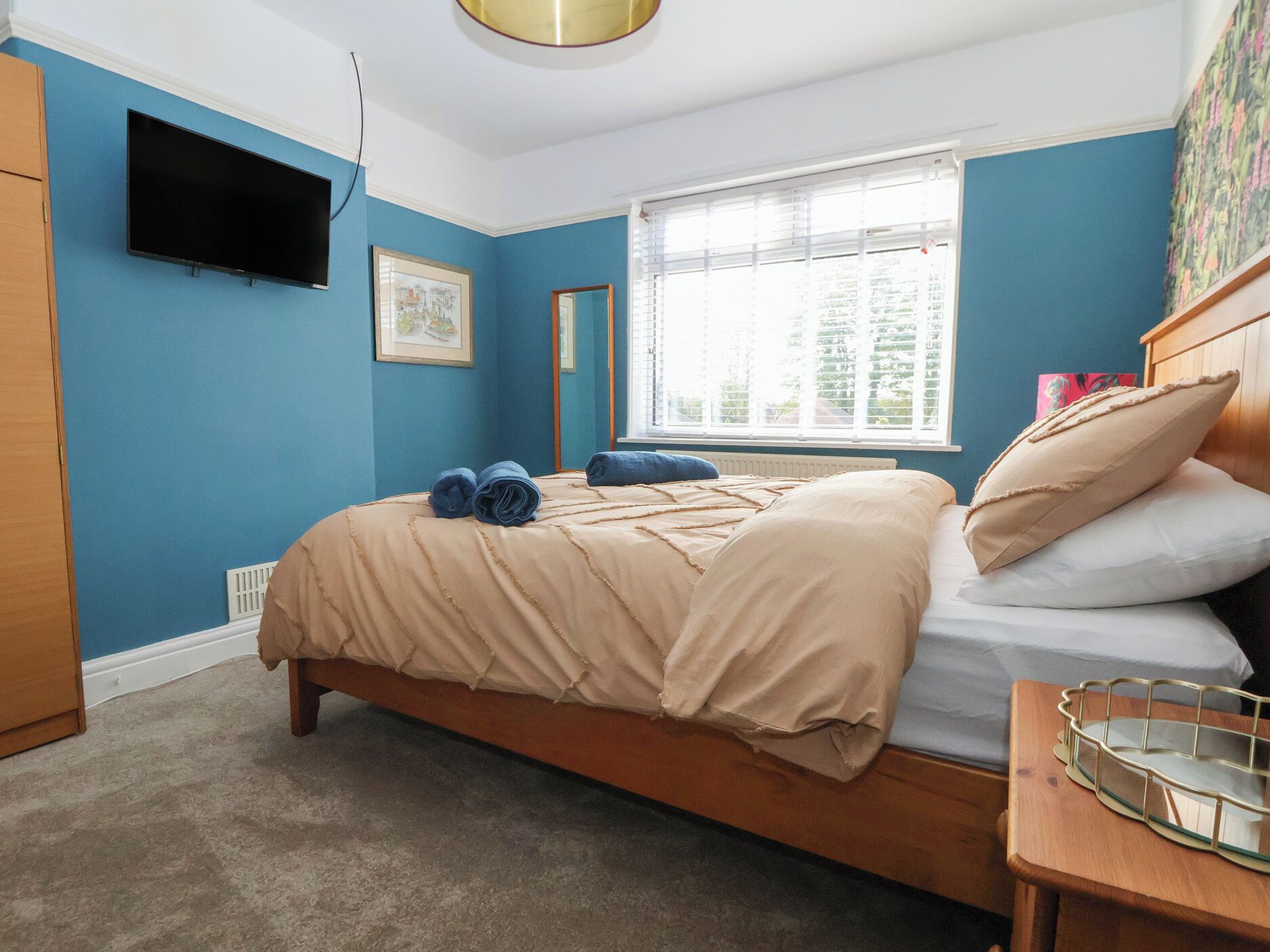 A bedroom with a bed, a bedside table, a TV mounted on the wall, and a window with blinds.
