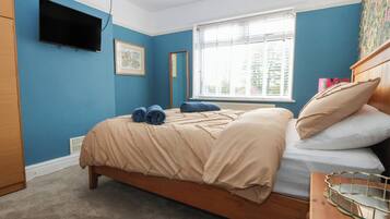 A bedroom with a bed, a bedside table, a TV mounted on the wall, and a window with blinds.