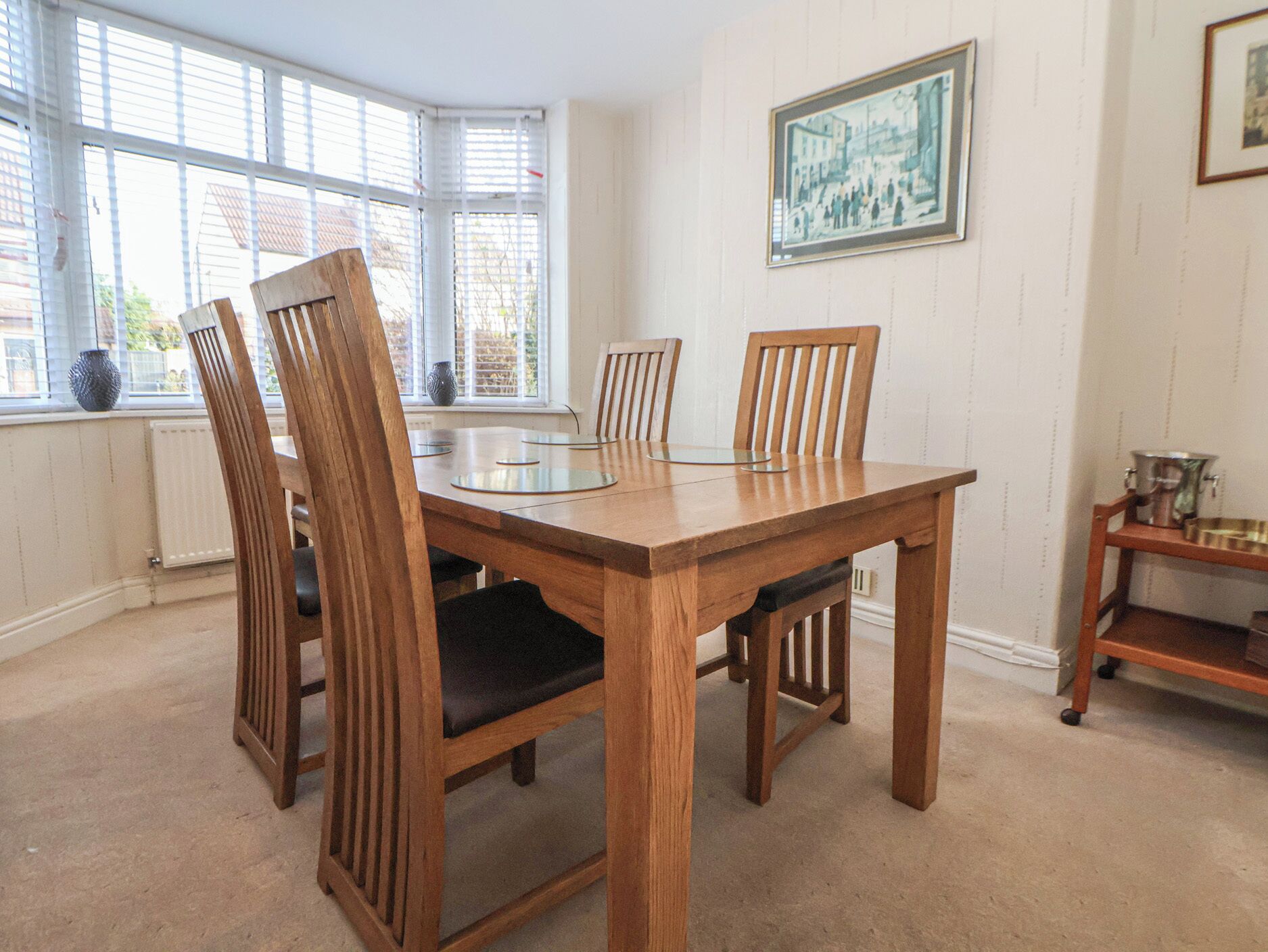 A dining area with a wooden table and chairs, large windows with blinds, and a framed picture on the wall.
