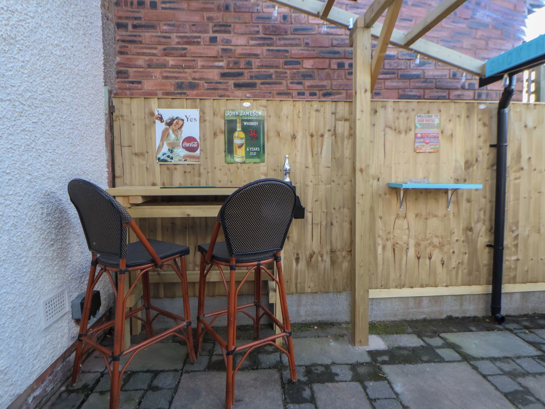 An outdoor seating area with two wicker chairs and a wooden table, a wooden fence, and signs advertising John Jameson whiskey and Coca-Cola.