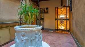 Un patio con una fuente de piedra blanca que presenta querubines, una planta en maceta y una reja de hierro decorativa con una luz cálida.