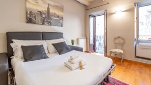 Una habitación de hotel con una cama grande, un cuadro de paisaje urbano, un radiador, una silla y una ventana con vistas.