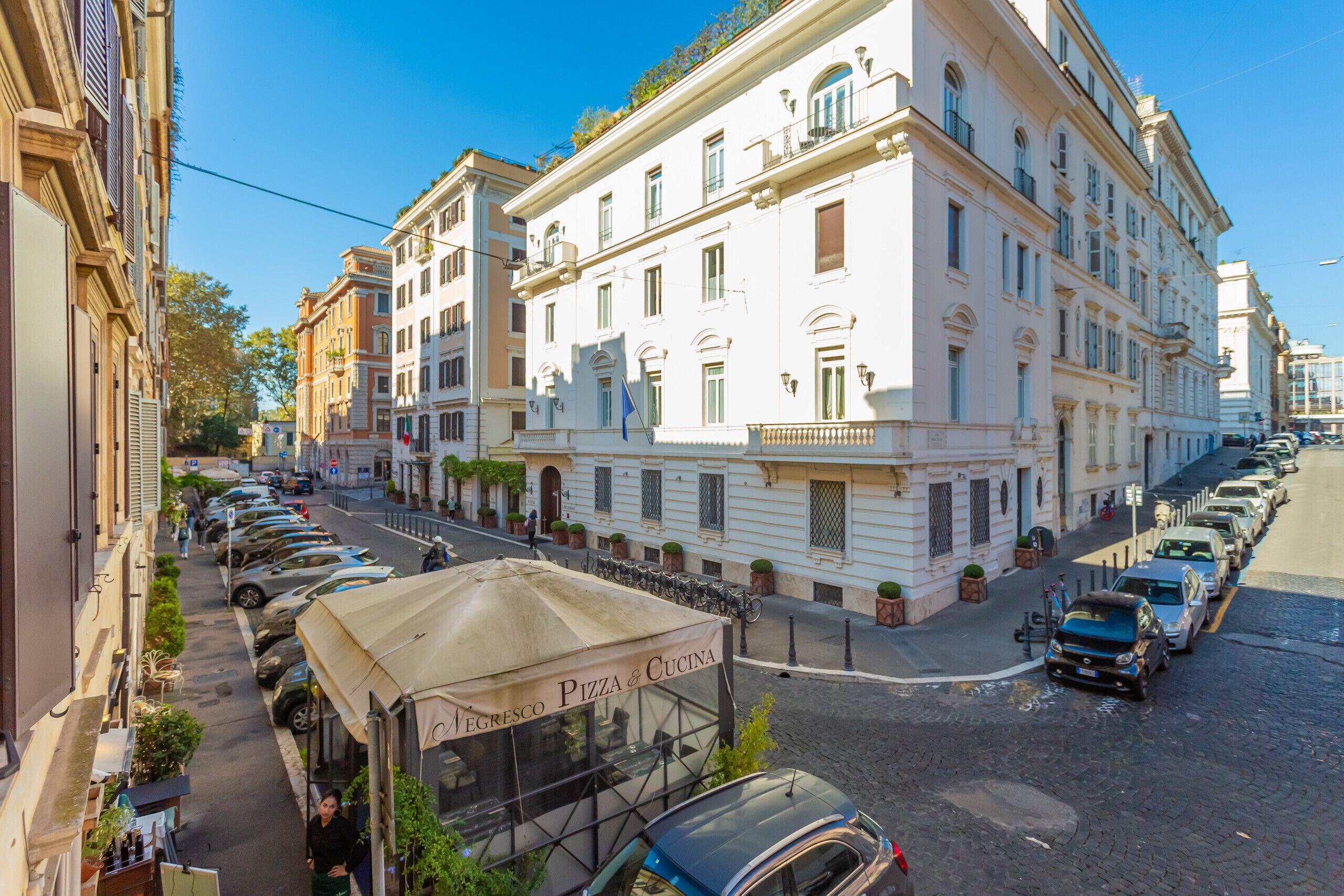 Une rue bordée de voitures garées, un immeuble avec une enseigne indiquant « PIZZA & CUOCINA », et un ciel bleu dégagé.