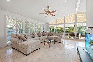 Ein geräumiges Wohnzimmer mit einem beigefarbenen Ecksofa, einem Glastisch und einem Deckenventilator. Große Fenster sorgen für natürliches Licht und einen Blick ins Freie.