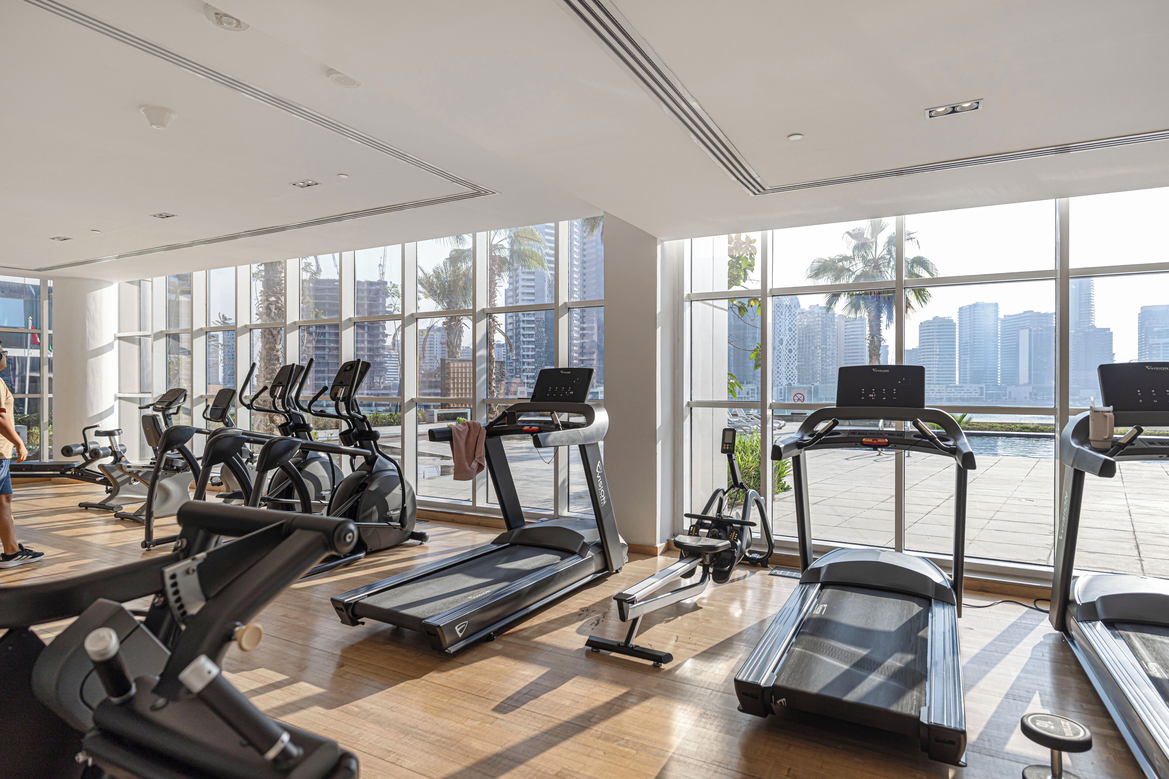A gym with multiple treadmills, exercise bikes, and a view of a cityscape through large windows.