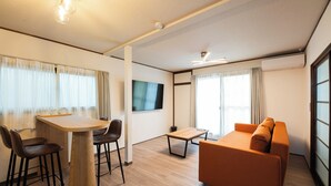 A modern living room with a wooden dining table, two bar stools, an orange sofa, and a flat-screen TV mounted on the wall.