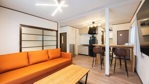 A modern apartment with a wooden floor, orange sofa, wooden table, kitchenette, and a ceiling light fixture.