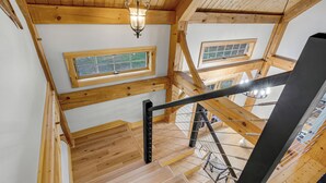 A wooden staircase with a metal handrail, wooden beams, and a ceiling light. - Timberframe Cabin with Hot Tub Close to Sunday River! (Bethel)