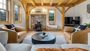 A cozy living room with wooden beams, a stone fireplace, a large TV, and comfortable seating. - Timberframe Cabin with Hot Tub Close to Sunday River! (Bethel)