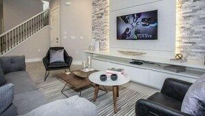 A modern living room with a grey sofa, a black armchair, a coffee table, and a TV mounted on a brick wall. - Summerville Resort 5 Bedroom Vacation Home 2620 (Kissimmee)
