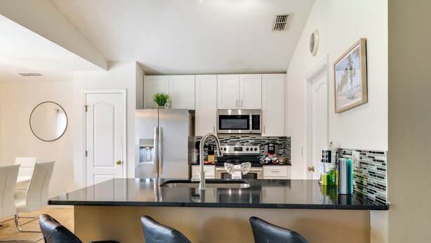 A modern kitchen with white cabinets, stainless steel appliances, a black countertop, and three black bar stools. - Peaceful Clermont Gem w/ Pool, 9 Mi to Disney! (Clermont)
