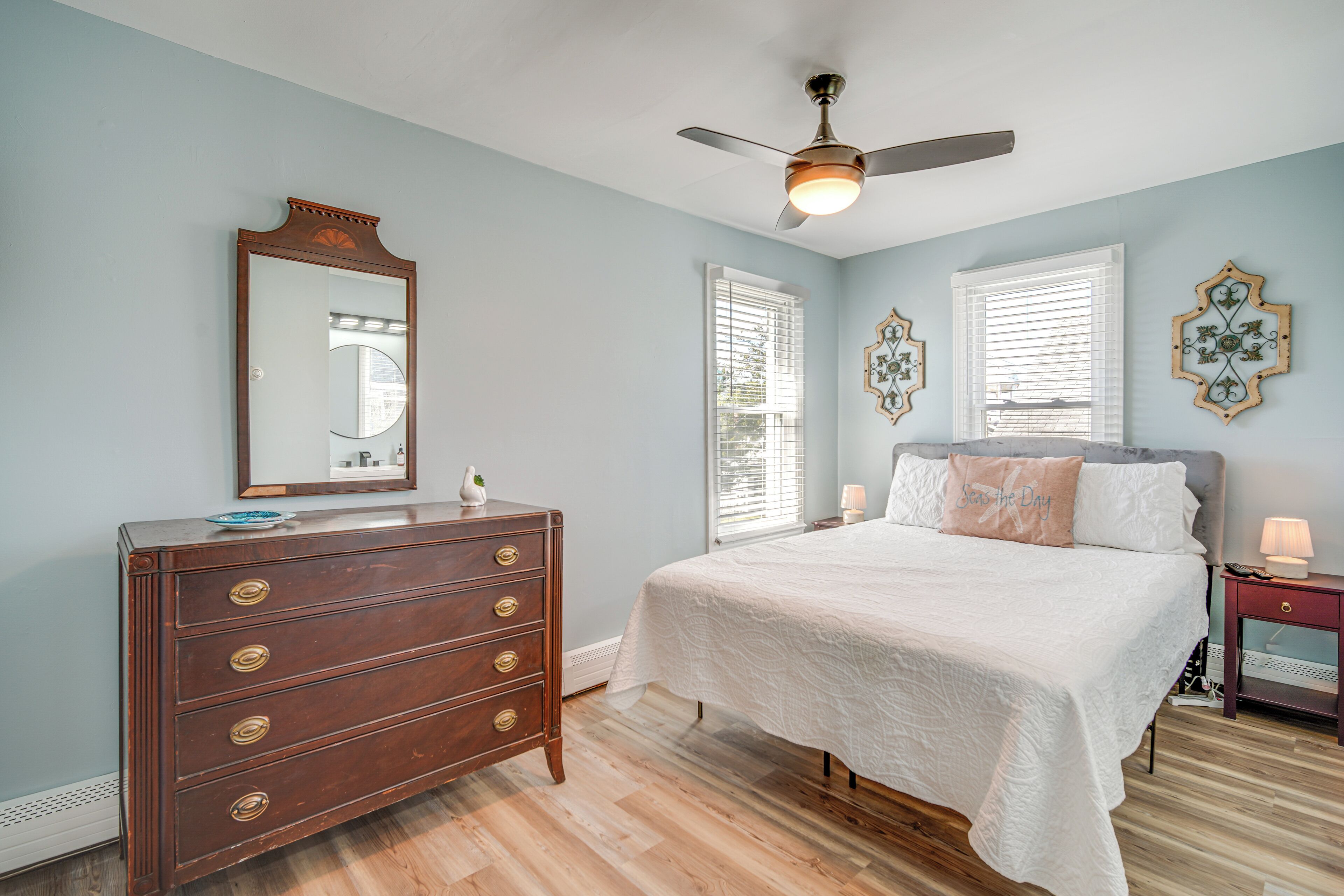 Un dormitorio con cómoda de madera, cama con ropa de cama blanca, ventilador de techo y decoración en la pared.