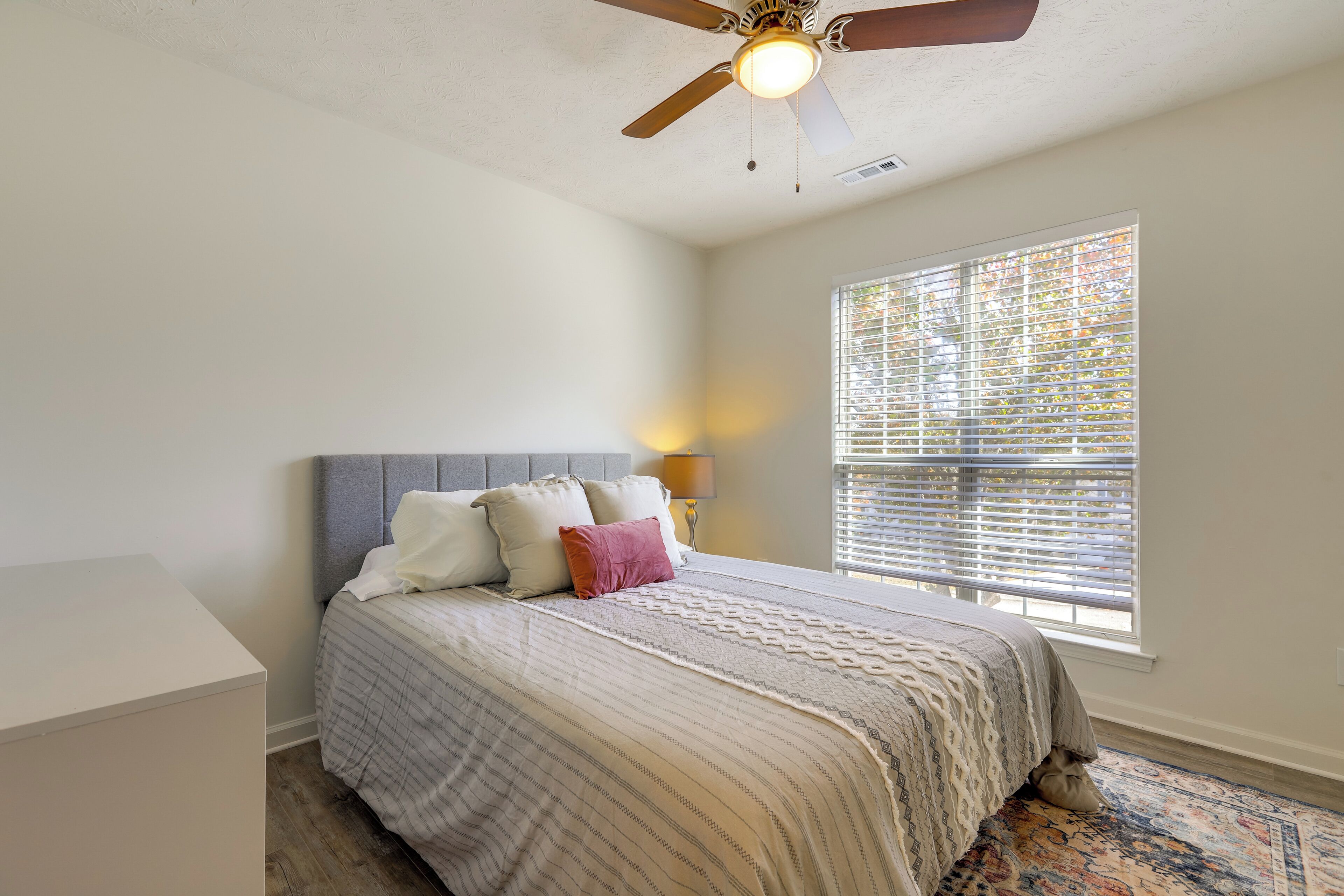 A bedroom with a bed, a ceiling fan, a window with blinds, and a nightstand with a lamp.