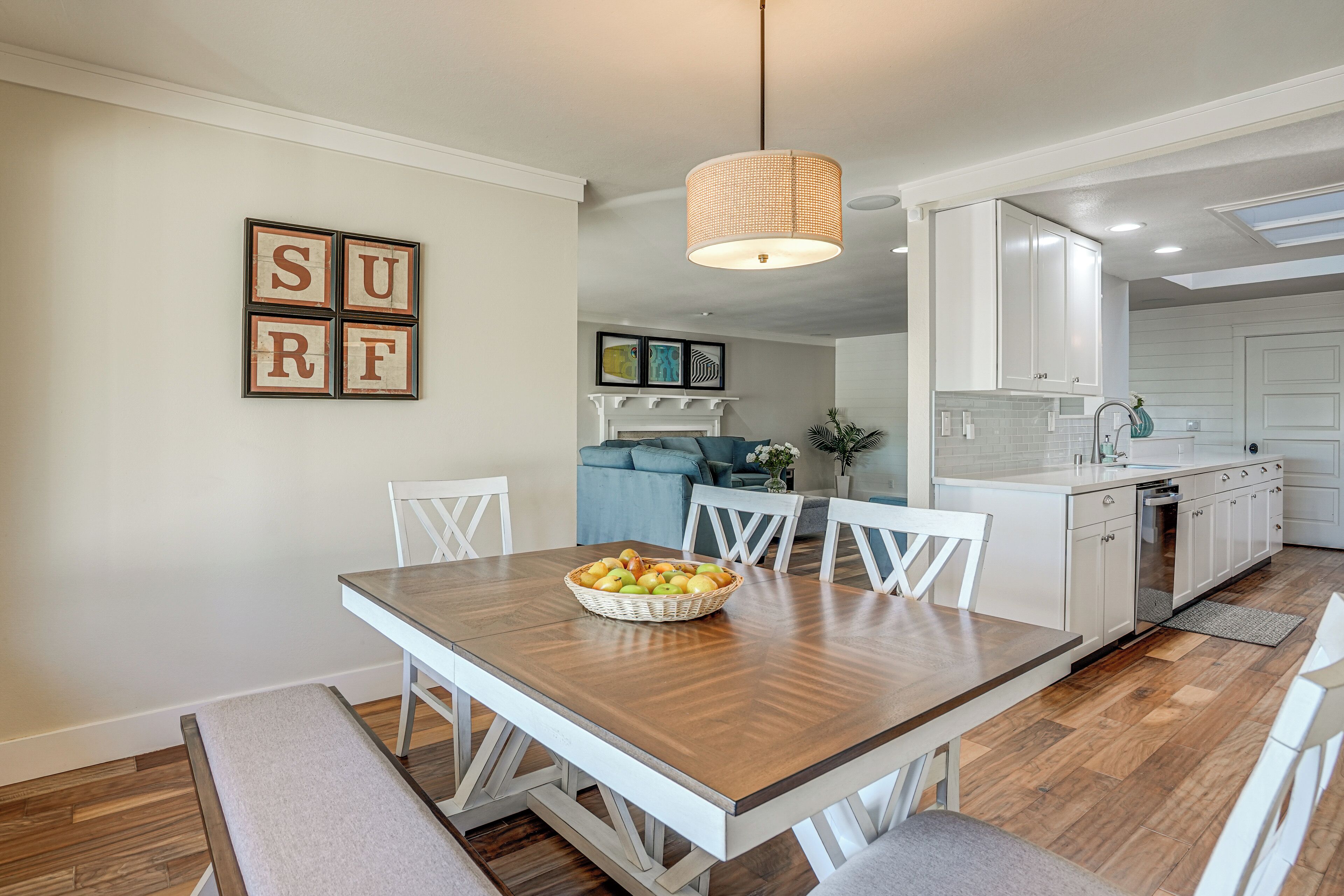 A dining area with a wooden table, white chairs, and a basket of fruit. There is a white cabinet and a kitchen in the background.