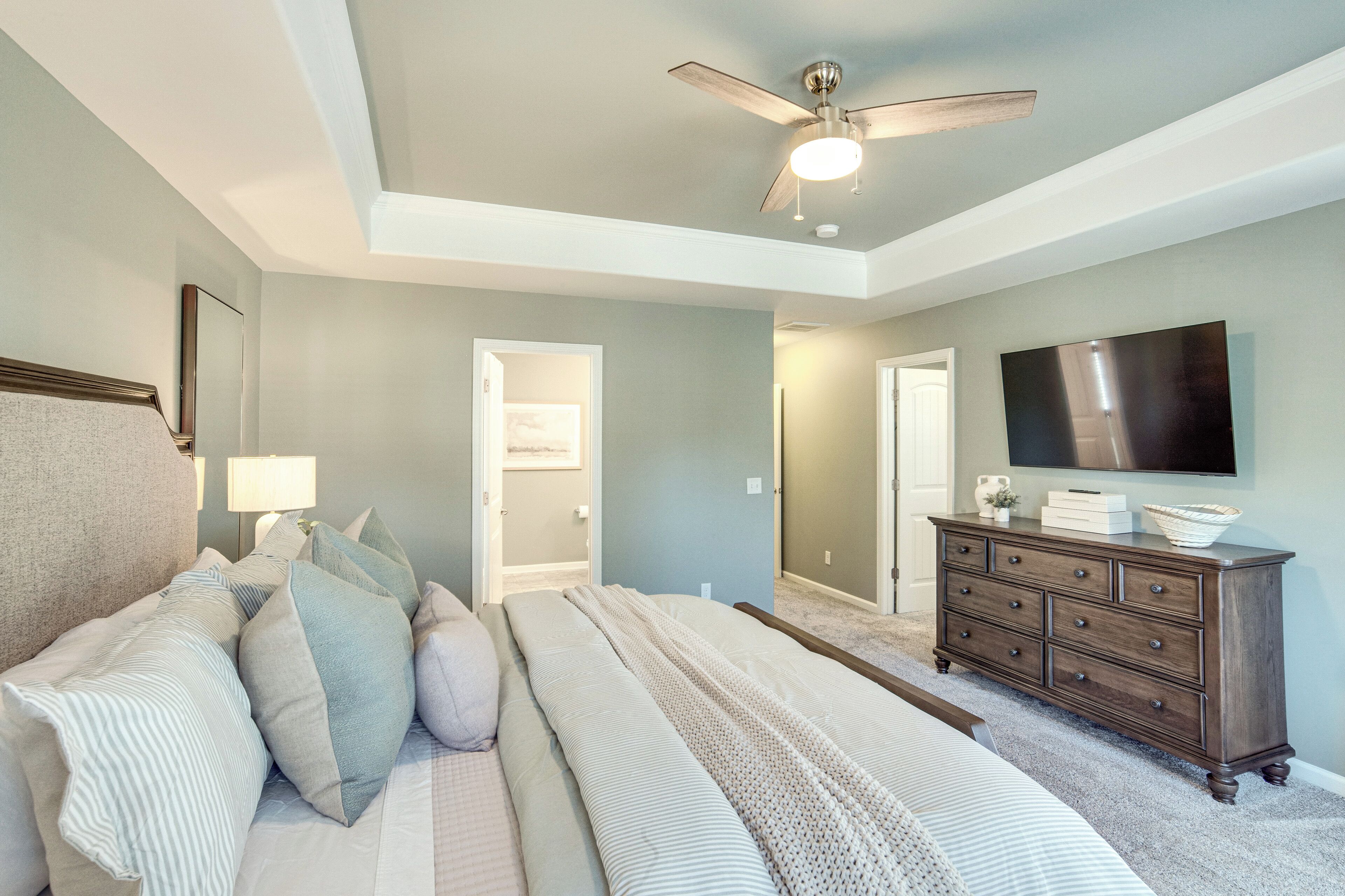 A bedroom with a bed, dresser, ceiling fan, and a TV on the wall.