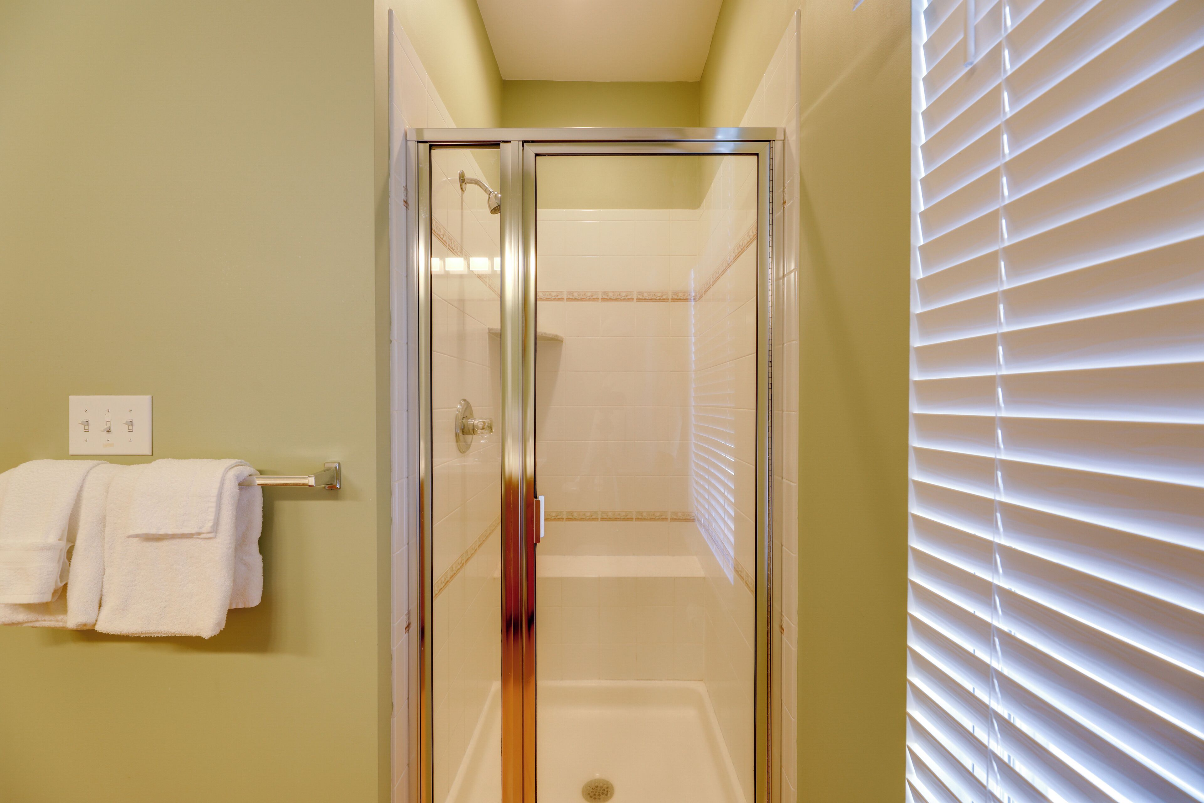 A glass-enclosed shower with a white bathtub, a towel rack with white towels, and a window with blinds.