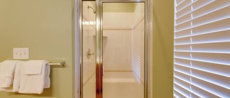 A glass-enclosed shower with a white bathtub, a towel rack with white towels, and a window with blinds.