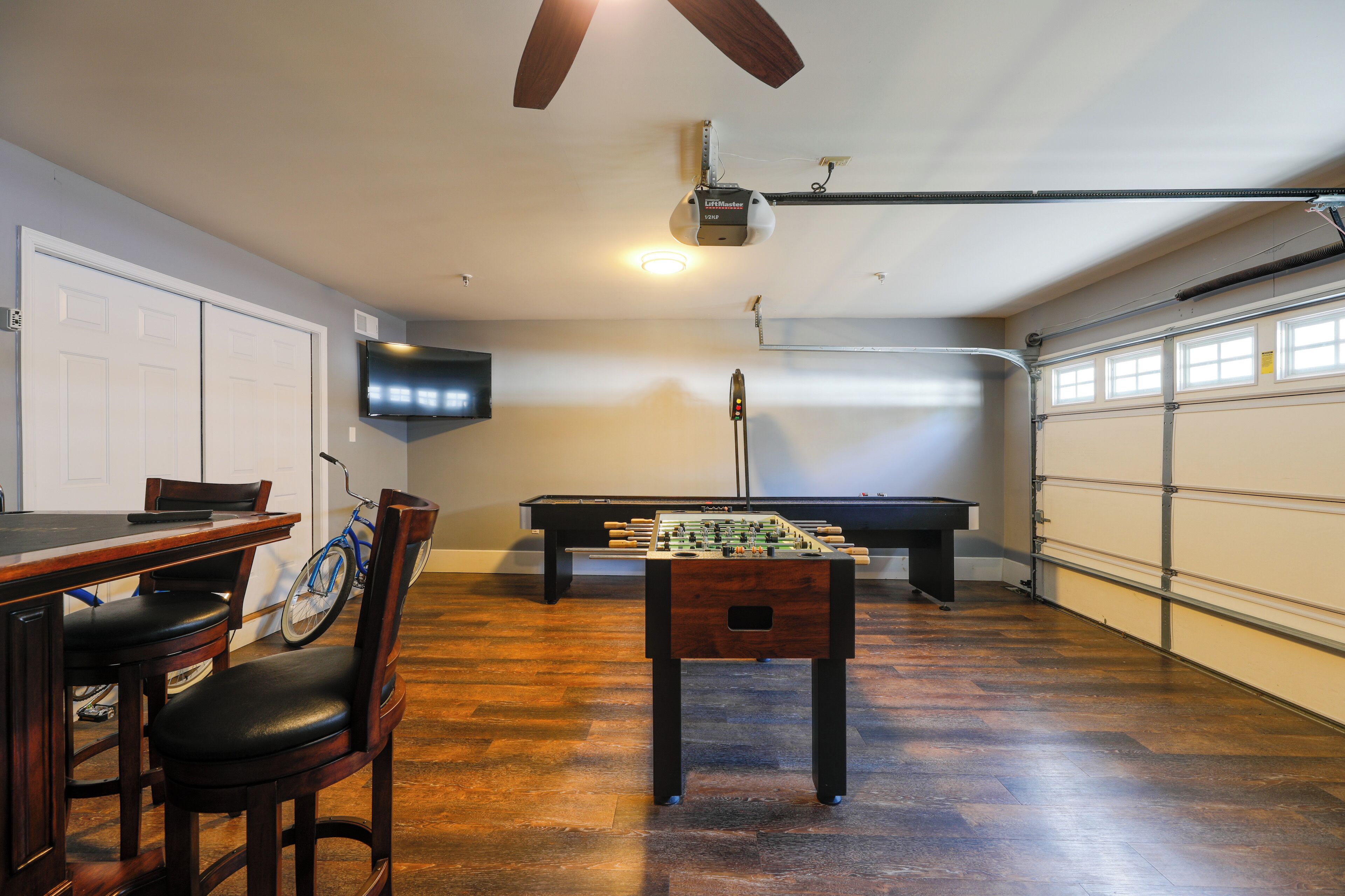 A room with a foosball table, a pool table, a TV mounted on the wall, and a ceiling fan.
