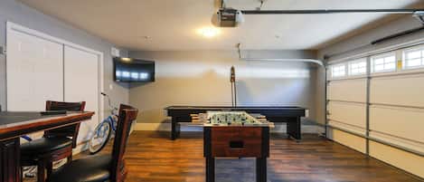 A room with a foosball table, a pool table, a TV mounted on the wall, and a ceiling fan.