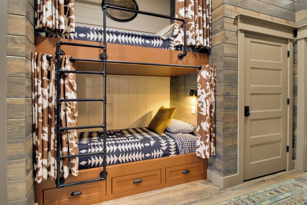 A bunk bed with a ladder, a wooden headboard, and a patterned bedspread. There is a door to the right and a patterned rug on the floor. - Fly Fishers Paradise: Getaway Near White River! (Cotter)