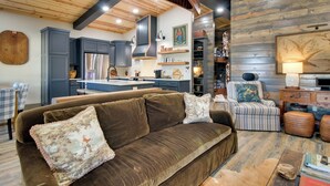 Un salon chaleureux avec un canapé marron, un fauteuil à rayures et un pouf. On peut voir un plafond et des murs en bois, ainsi que des étagères intégrées et une cheminée.