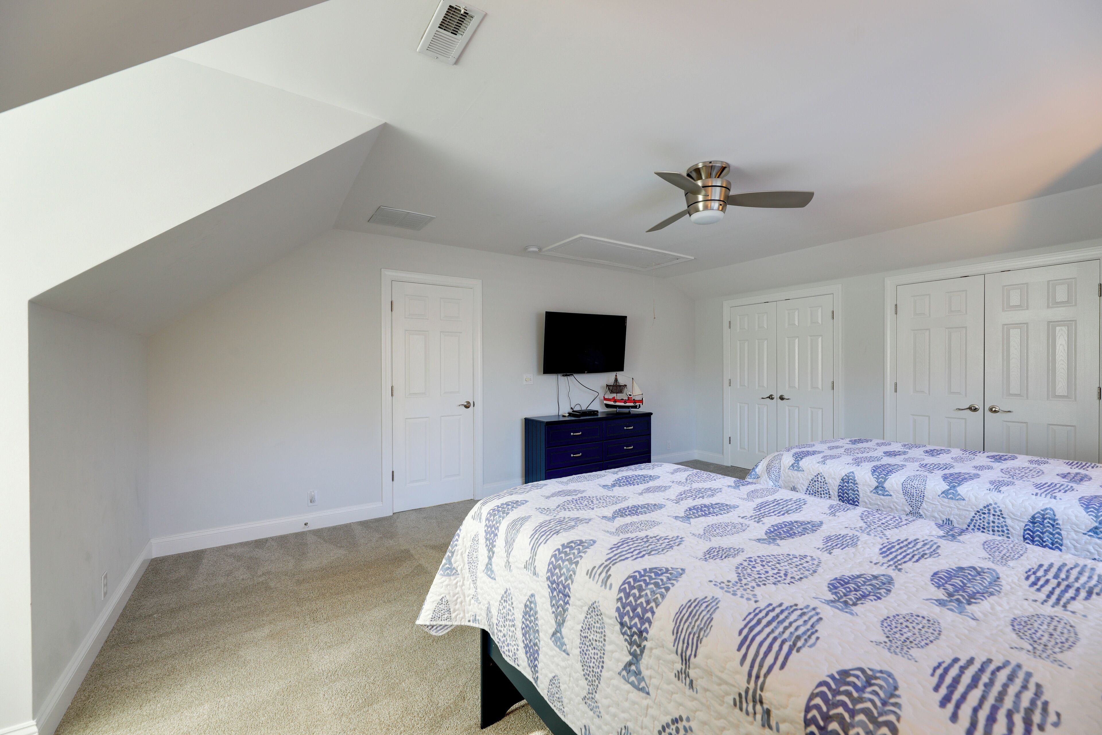 Une chambre avec un plafond en pente, un ventilateur de plafond, un lit recouvert d’une couvre-lit à motifs, une commode et un téléviseur fixé au mur.