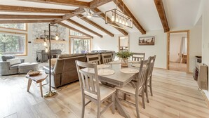Ein geräumiger Loft mit freiliegenden Holzbalken, einem Essbereich mit Holzsesseln und einem Tisch, einem gemütlichen Wohnzimmer mit einer Couch und Kamin, sowie großen Fenstern mit Blick ins Freie.
