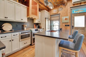 A kitchen with white cabinets, stainless steel appliances, a wooden ceiling, and a large island with blue chairs. - Walk to White River: Cozy Escape w/ Screened Porch (Cotter)