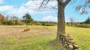 Un arbre à la base duquel se trouve une pile de bûches, une construction en briques en arrière-plan et un champ herbeux parsemé de feuilles.