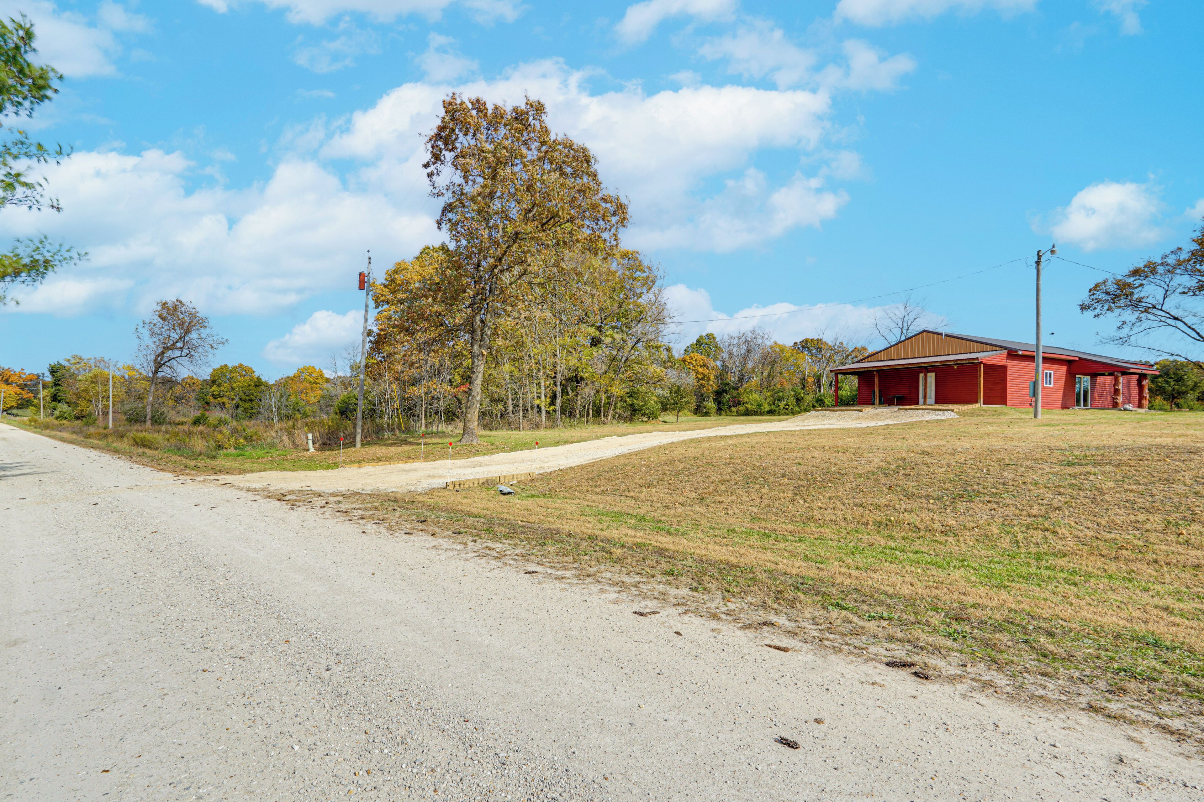 A rural road leading to a red building with a sloped roof, surrounded by trees and a grassy area.