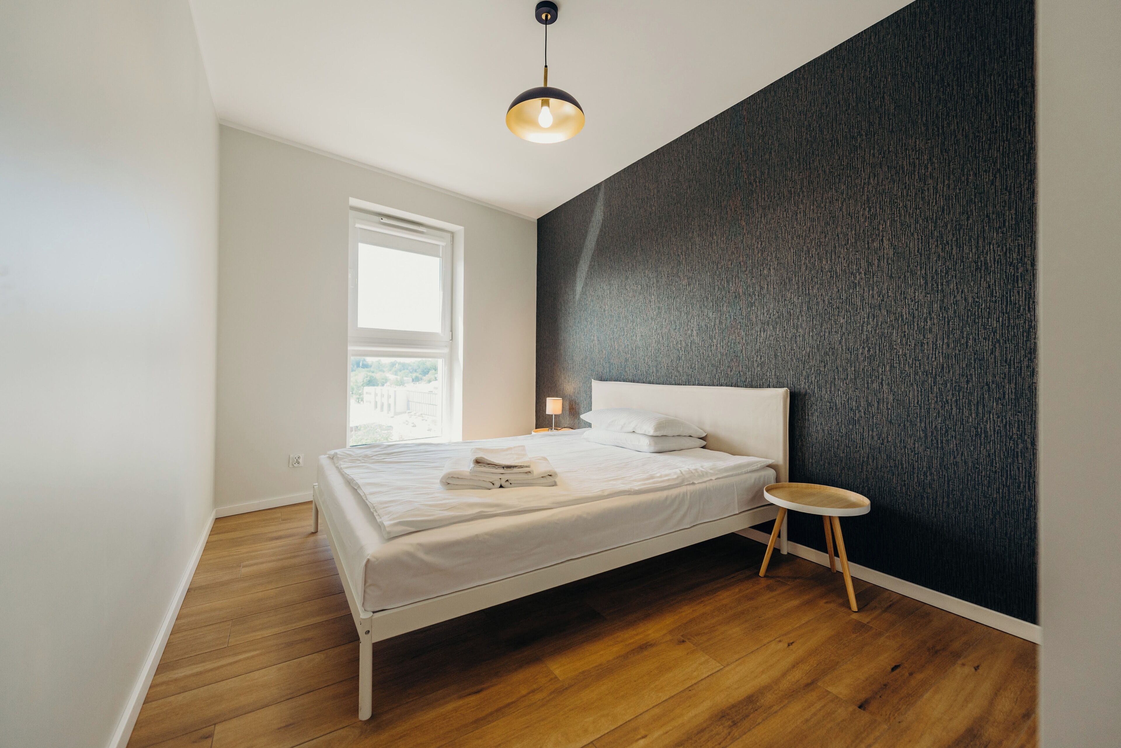 A modern bedroom with a bed, a small round table, a window, and a textured wall.