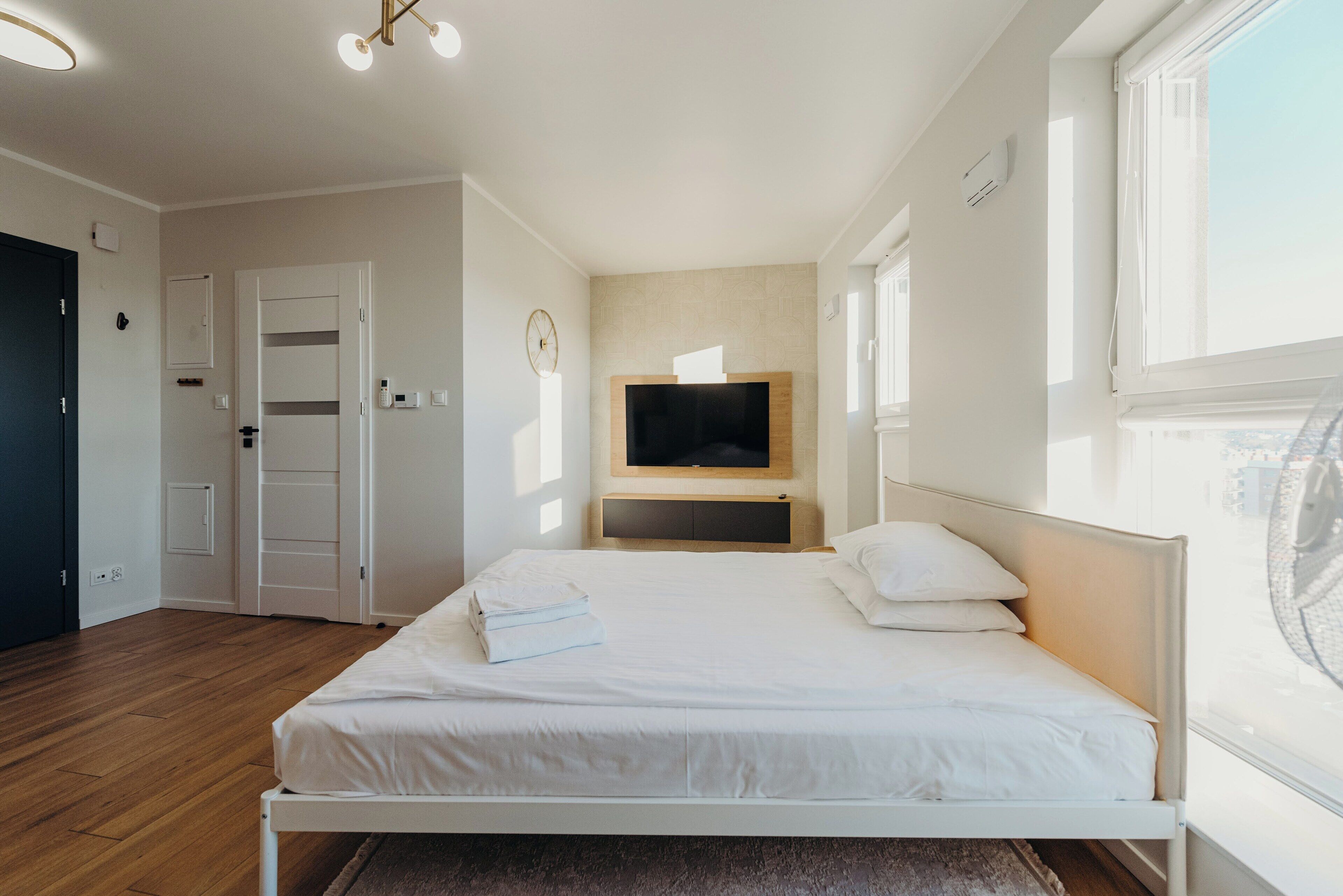 Ein modernes Schlafzimmer mit Holzboden, einem Bett mit weißen Leinen, einem an der Wand montierten Flachbildfernseher und einem Fenster, das für natürliches Licht sorgt.