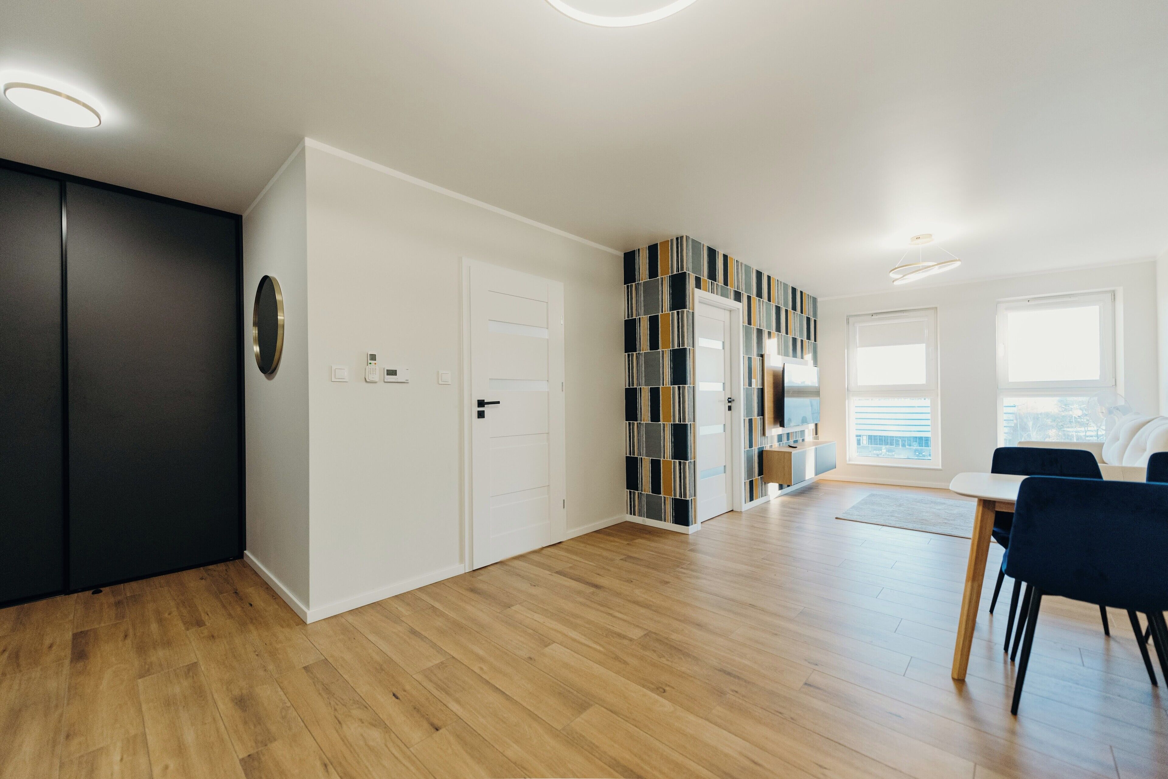 A modern living room with a wooden floor, a black wardrobe, a white door, and a dining area with a table and chairs.