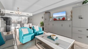 A modern kitchen with a dining area, a living room with blue sofas, a TV, and a large grey cabinet. - Rising Tides Properties Condo 1007 (Murrells Inlet)