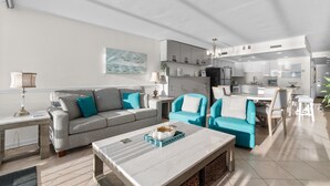 A modern living room with a grey sofa, blue armchairs, and a marble coffee table. A kitchen with white cabinets and a dining area with a table and chairs are visible. - Rising Tides Properties Condo 1007 (Murrells Inlet)