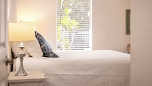 A neatly made bed with a white bedspread, a bedside lamp, and a window with blinds.
