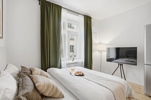 A hotel room with a bed, a TV on a stand, a window with curtains, and a bedside table with a glass of orange juice and a plate of food. - Nordic Deluxe / Renovated / Central - A/C - WiFi (Helsinki)