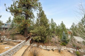 A landscaped garden with a stone wall, a wooden deck, and various trees. - Big Bear Nest at Eagle Mountain - Game Room, Pool Table, Ping Pong, Foosball, Jet Tub, 3 Fireplaces (Big Bear Lake)