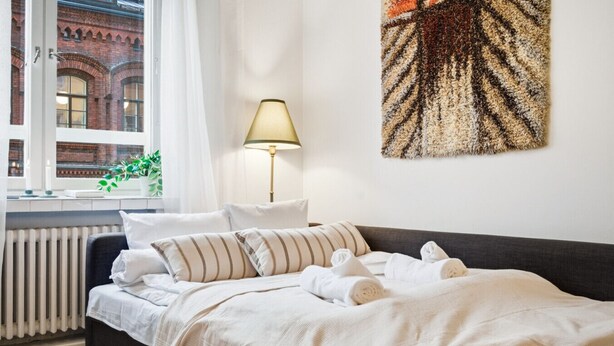 Ein ordentlich bezogenes Bett mit gestreiften Kissen und aufgerollten Handtüchern, eine Nachttischlampe und Blick auf ein rotes Backsteingebäude durch ein Fenster.