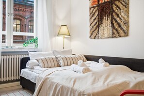 En pænt redt seng med stribede puder og sammenrullet håndklæder, en senget lampe og udsigt til en murstensbygning gennem et vindue.