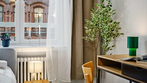 Ein Hotelzimmer mit Bett, Nachttisch mit Lampe, Schreibtisch mit Stuhl, Zimmerpflanze und Blick auf ein Backsteingebäude durch ein Fenster mit durchsichtigen Vorhängen.