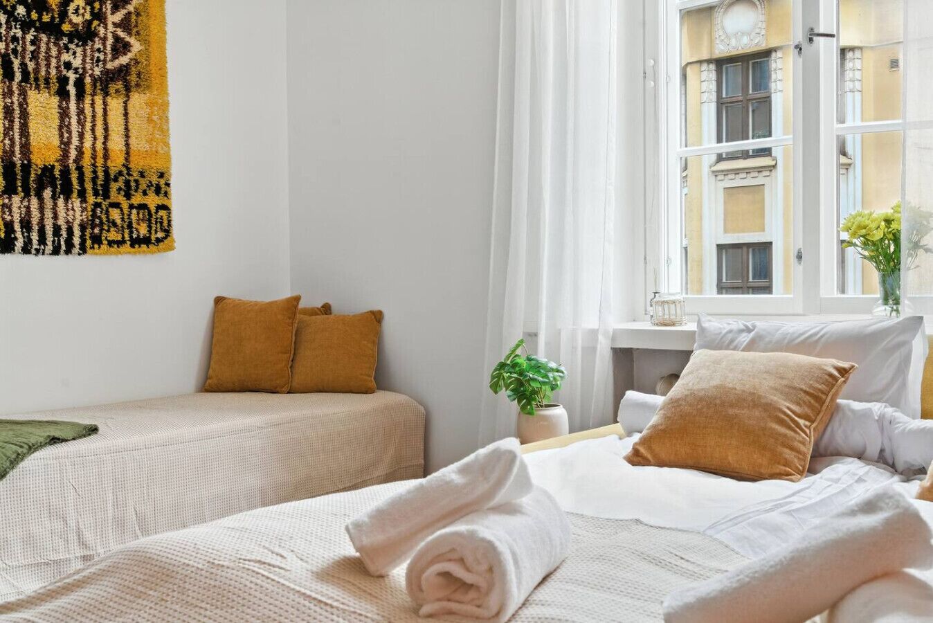 Une chambre d’hôtel avec deux lits, une fenêtre donnant sur des immeubles, une plante posée sur une table d’appoint et un tableau accroché au mur.