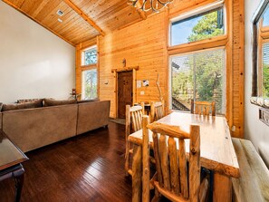 A wooden interior with a dining table and chairs, a sofa, and large windows. - Bear State of Mind - Close to Ski Slopes, Hot Tub, Foosball, Fireplace, Deck, BBQ (Big Bear City)